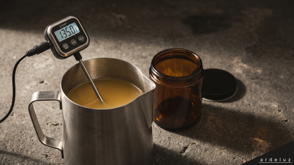 Calibrated probe thermometer reading 135F in a pitcher of melted soy wax beside an amber candle vessel on dark concrete, illustrating candle pour temperature.